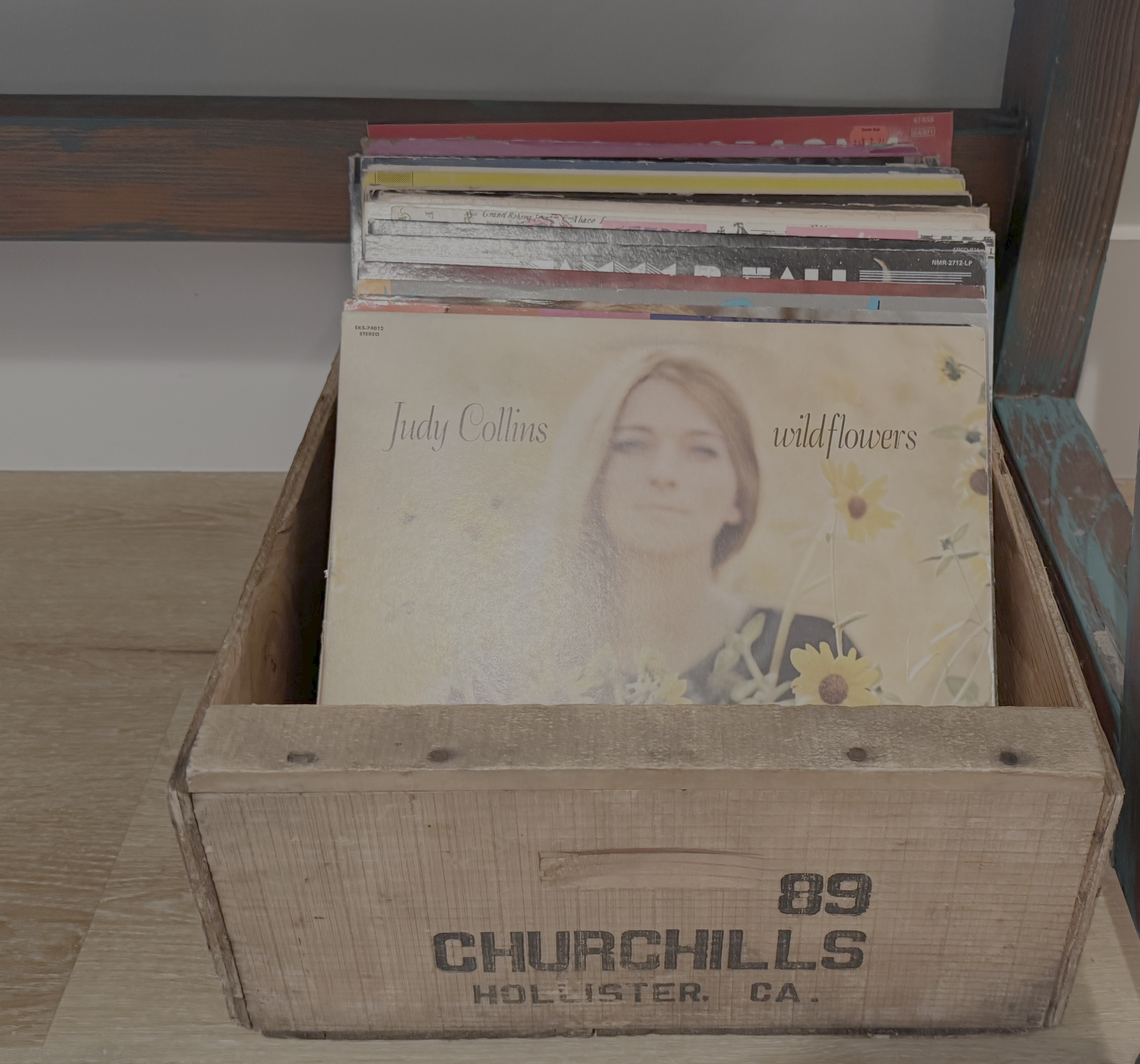 Auto-generated description: A wooden crate labeled CHURCHILLS HOLLISTER, CA. holds a collection of vinyl records, with Judy Collins' Wildflowers album prominently displayed at the front.